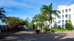 A paved pathway lined with palm trees and a white building with multiple windows and balconies. - Stay.Plus Almasi Beachfront Apartments (Mombasa)