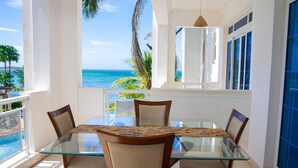 A dining area with a glass table and chairs, a view of the ocean, and a balcony with a railing. - Stay.Plus Almasi Beachfront Apartments (Mombasa)