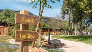 A wooden signpost with 'Accueil' and 'Les Gites' written on it, set against a backdrop of a clear sky, trees, and a pathway.