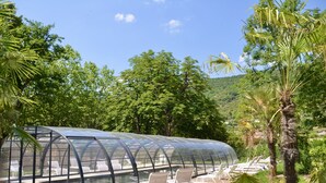 A glass-enclosed lounge area with white chairs and a pool, surrounded by lush greenery and trees.