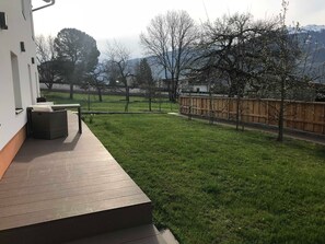 Eine Terrasse mit Holzboden, einer Rasenfläche und Blick auf Bäume und Berge.