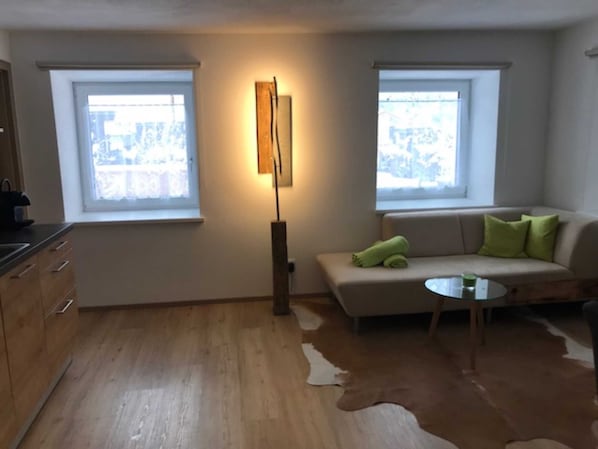 Un salon moderne avec un canapé, une petite table ronde et un tapis en peau de vache. Il y a deux fenêtres équipées de stores, une lampe sur pied et un coin cuisine avec des meubles en bois.