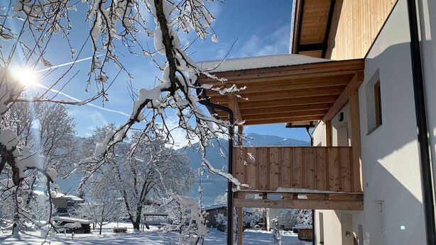 Lumipeitteinen ulkoalue, jossa on puinen pergola, parvekkeellinen rakennus ja kirkas taivas.