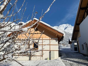 Un chalet en bois, dont le toit est recouvert de neige, avec une enseigne indiquant « Schusterbrand », et une chaîne de montagnes en arrière-plan.