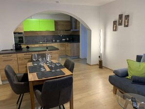 A modern kitchen with green cabinets, a dining area with a wooden table and chairs, and a living area with a sofa and a glass coffee table. - Gravensteiner - Schusterbrand Apartments (Absam)