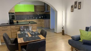 A modern kitchen with green cabinets, a dining area with a wooden table and chairs, and a living area with a sofa and a glass coffee table. - Gravensteiner - Schusterbrand Apartments (Absam)