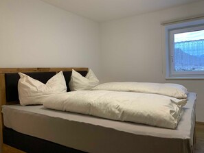 Un lit simple avec une tête de lit en bois, une literie blanche et une fenêtre donnant sur un paysage enneigé.