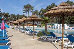 Área de piscina com espreguiçadeiras e guarda-sóis de palha, cercada por árvores e um edifício com fachada azul e branca.