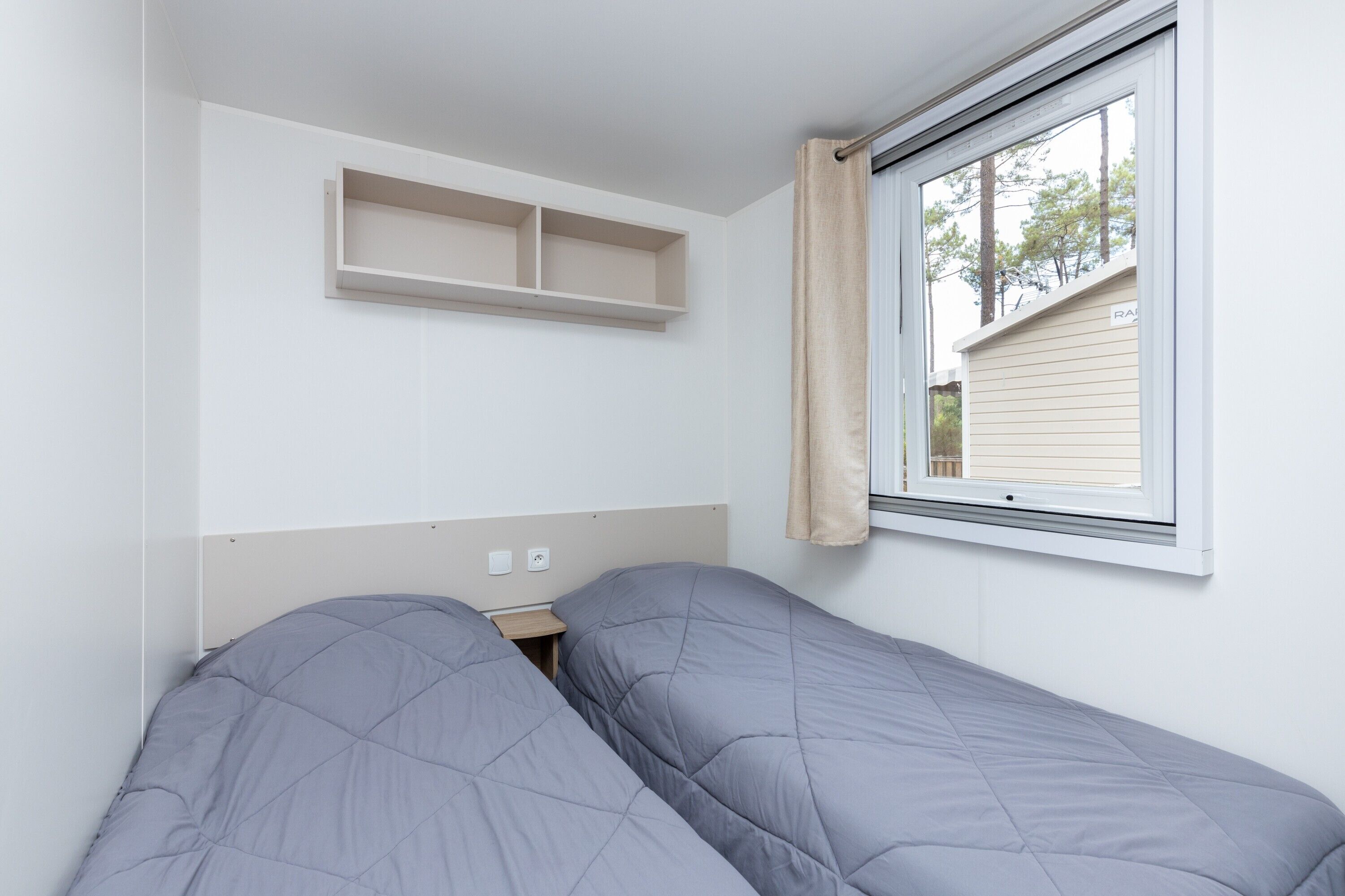 Une chambre avec deux lits simples, une fenêtre donnant sur des arbres et un mur blanc avec une étagère.