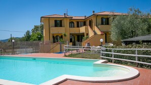 Une piscine avec une terrasse carrelée, un bâtiment à deux étages surmonté d’un toit en tuiles rouges, et un espace clos aménagé avec du mobilier d’extérieur.
