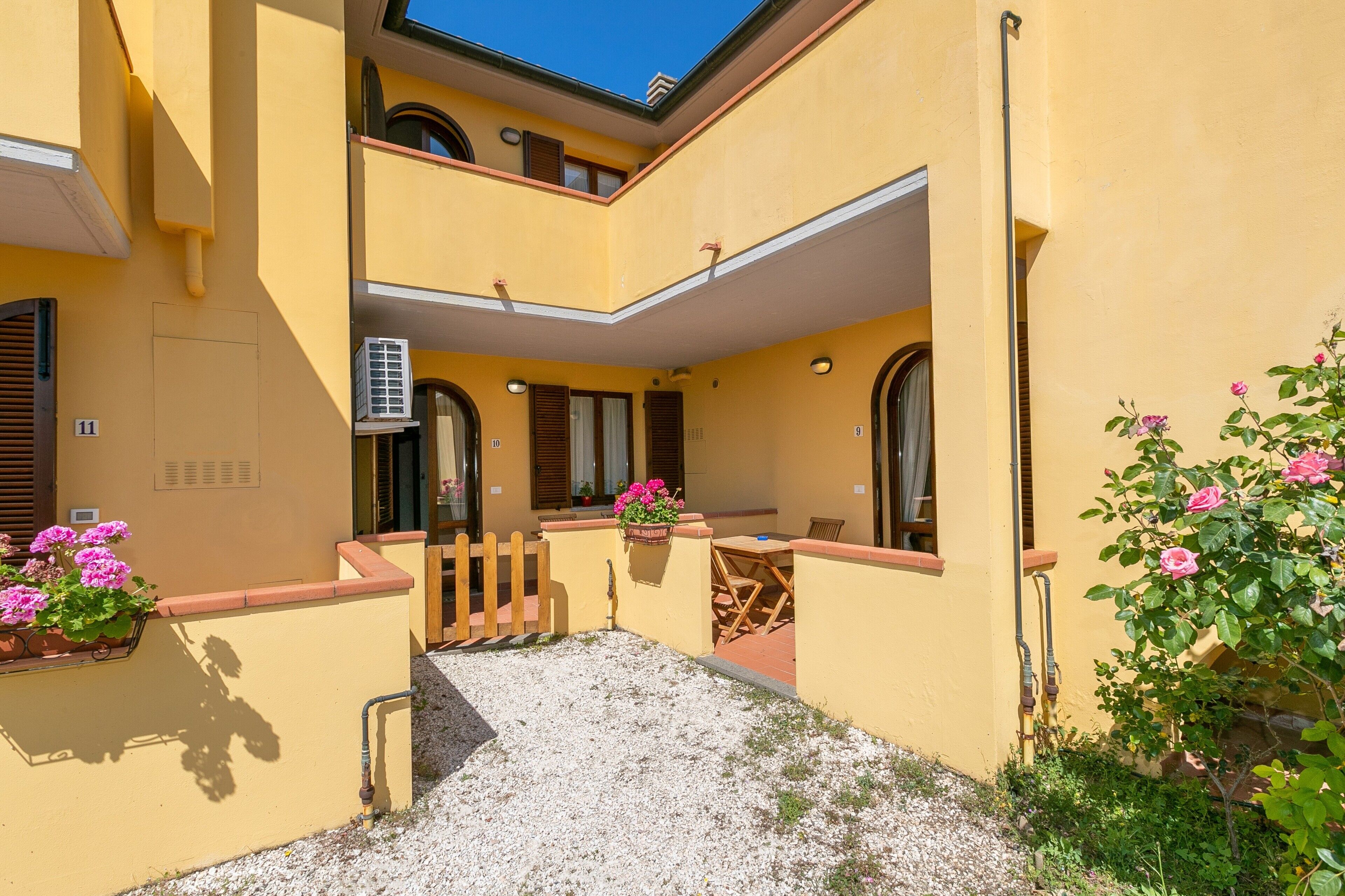 Un edificio giallo con una zona relax all'aperto coperta, un vialetto in ghiaia e piante in vaso.