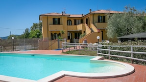 Une piscine avec une terrasse carrelée, un bâtiment à deux étages surmonté d’un toit en tuiles rouges et un espace clos aménagé avec du mobilier d’extérieur.