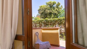 A room with a wicker chair, a wooden door, and a view of a garden with a tree and a hedge. - Apartment 'Bilocale Ciliegiolo' with Terrace, Shared Pool & Wi-Fi (Suvereto)