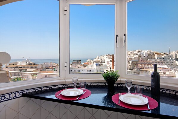 A dining area with a view of a coastal city, a potted plant, and a bottle of wine on the table.