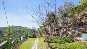 Uma trilha de pedra leva a um muro de pedra com uma porta verde, cercada por vegetação e um céu claro.