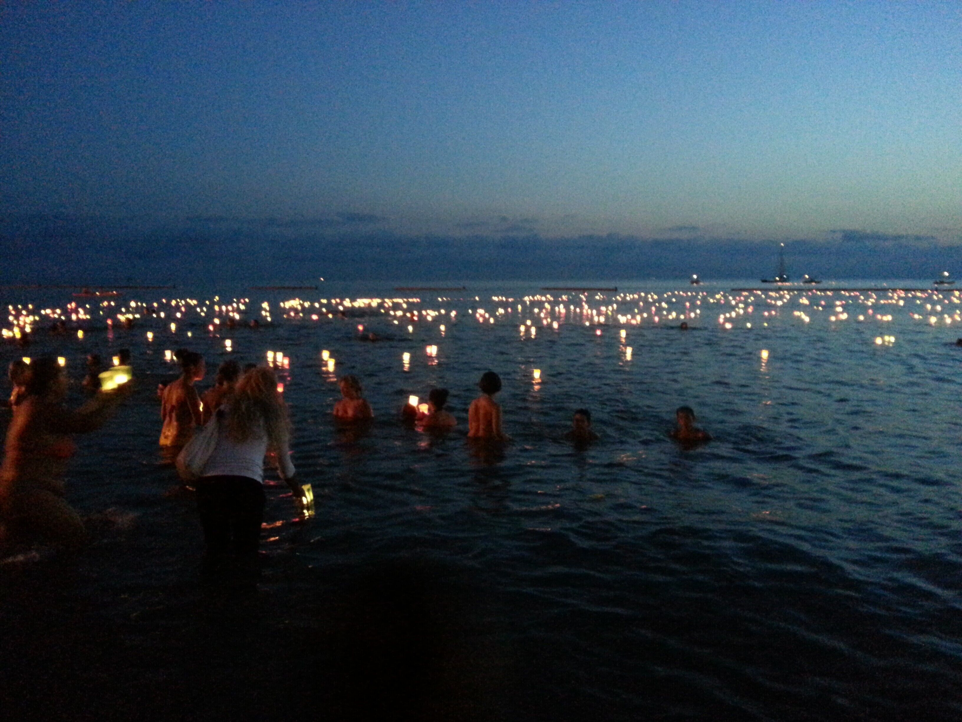 Escena nocturna con gente en el agua y una estructura iluminada a lo lejos.