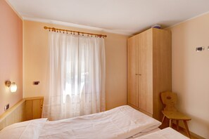 A bedroom with a bed, a wooden wardrobe, a wooden chair, and a window with sheer curtains.