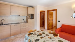 Una cucina compatta con elettrodomestici moderni, un'area pranzo con un tavolo apparecchiato per due e un accogliente divano rosso in un piccolo soggiorno.