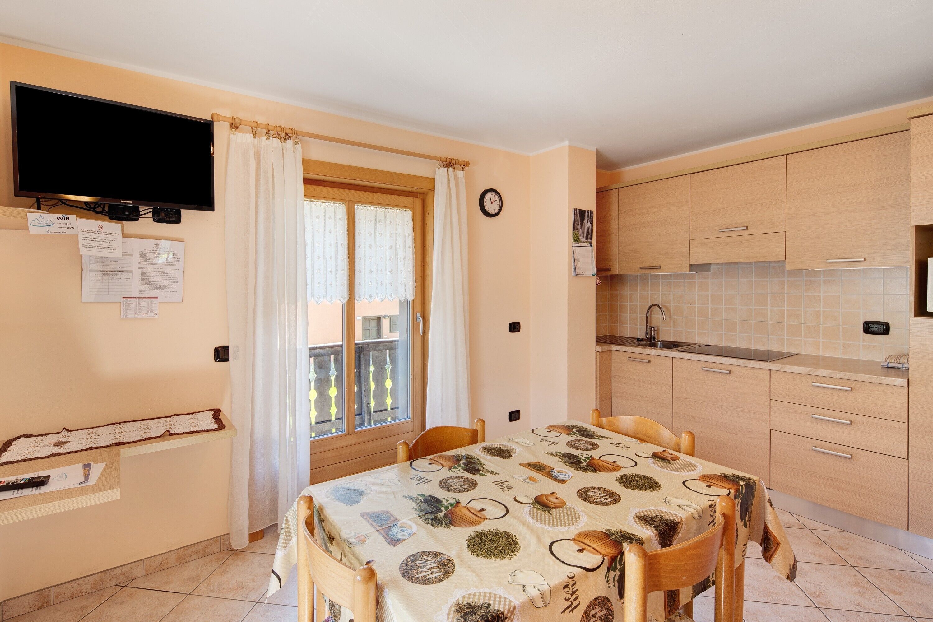 Een eetgedeelte met een tafel voor vier personen, een keuken met houten keukenkastjes, een spoelbak en een televisie boven het raam.