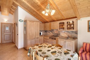 Une cuisine avec des meubles de rangement en bois, une table à manger recouverte d’une nappe et un lustre.