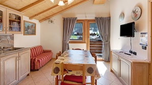 Un comedor con mesa de madera, sillas, un sofá, una cocina con armarios, un lavamanos, un televisor y una ventana con cortinas.
