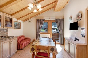 Un comedor con mesa de madera, sillas, un sofá, una cocina con armarios, un lavamanos, un televisor y una ventana con cortinas.