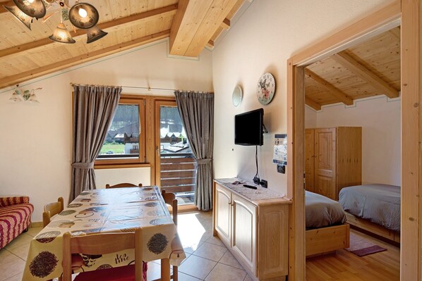 A room with a wooden ceiling, a dining table with chairs, a bed with a TV, and a window with curtains.