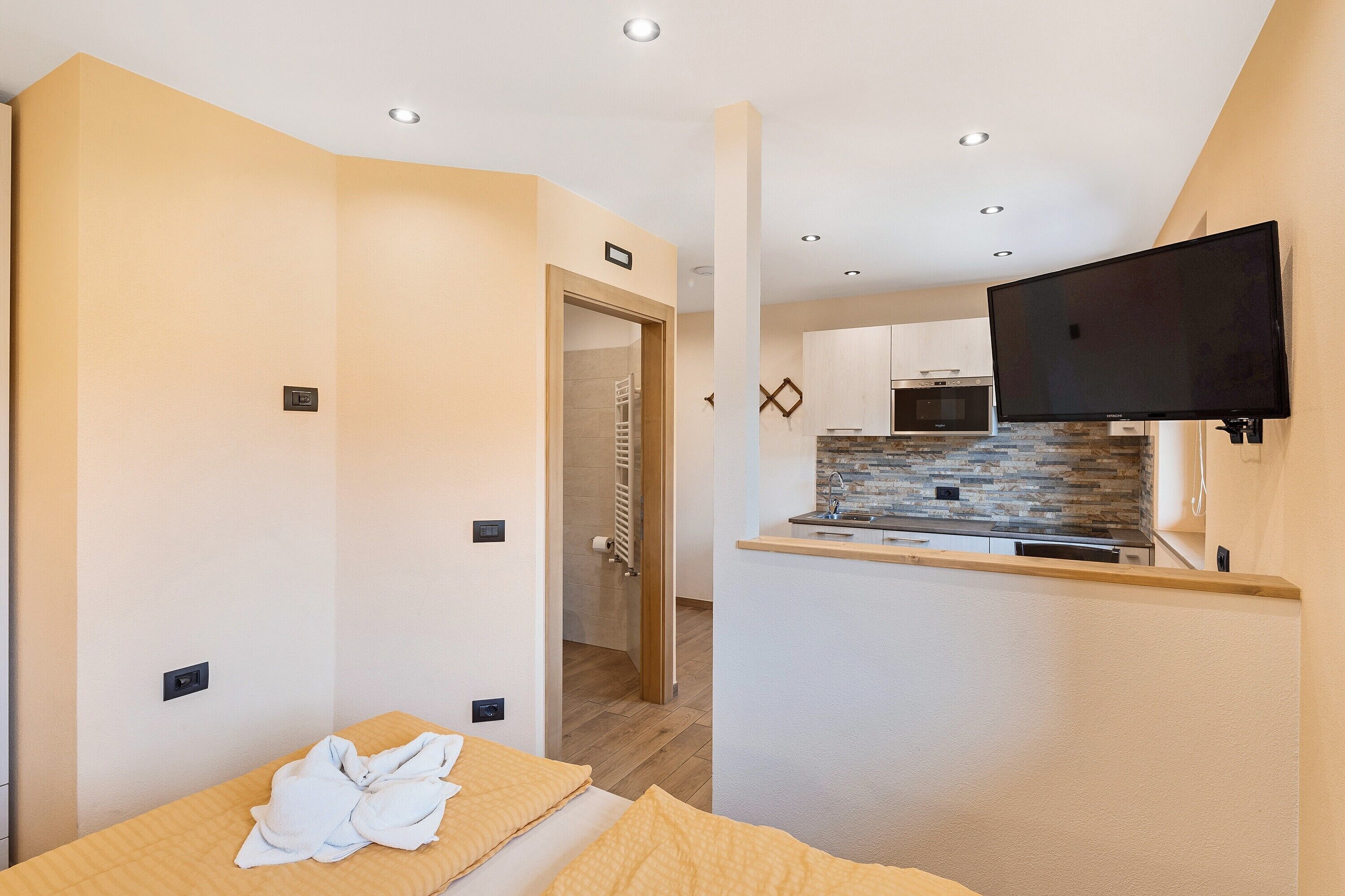 Ein modernes Hotelzimmer mit Bett, einem kleinen Tisch mit Handtüchern, einer Küchenzeile und einem an der Wand montierten Fernseher.