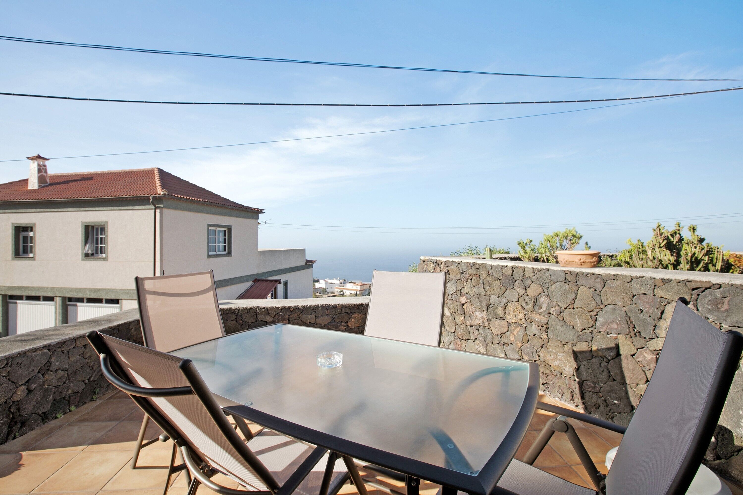 Une table à plateau en verre, entourée de chaises, sur un balcon, d’où l’on peut admirer un mur de pierre et un bâtiment avec un toit rouge.