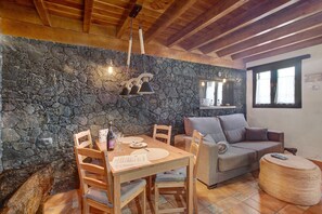 Ein Zimmer mit Steinwand, Holzbalkendecke, einem Esstisch mit Stühlen, einer Couch und einem runden Ottomanen.