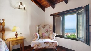 Una habitación con suelo de madera, un sillón floral, una mesita de noche con lámpara y una ventana con vistas al mar.