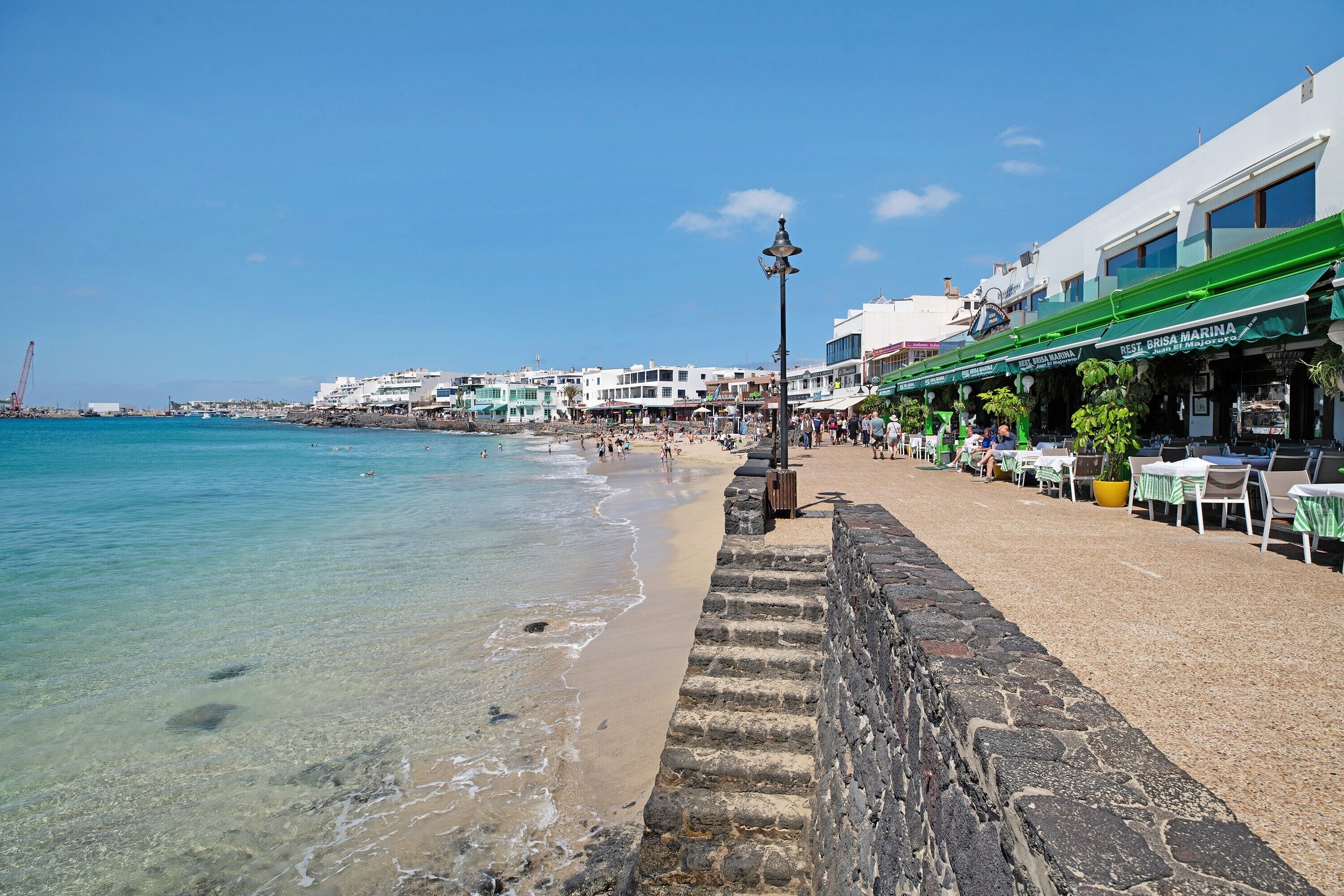 Un paseo marítimo con muro de piedra, playa y edificios frente al mar.