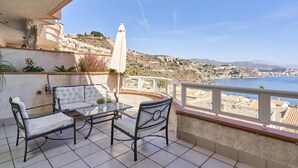 A balcony with outdoor furniture, a view of the sea, and a hillside in the background.