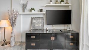 A modern living room with a flat-screen TV, a dark wooden cabinet with drawers, a lamp, decorative plants, and a 'Happy Home' sign.