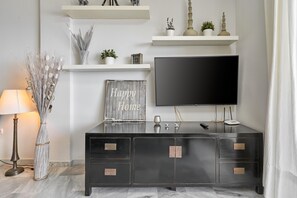 Un salon moderne avec un téléviseur à écran plat, un meuble en bois foncé doté de tiroirs, une lampe, des plantes décoratives et une pancarte portant l’inscription « Chez-nous, c’est le bonheur ».
