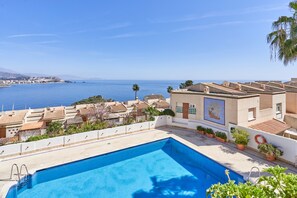 Une piscine sur le toit offrant une vue sur la mer, un bâtiment blanc orné d’une fresque murale et, au loin, des villas aux toits de tuiles rouges.