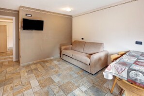 A room with a beige sofa, a television mounted on the wall, and a table with a patterned tablecloth.