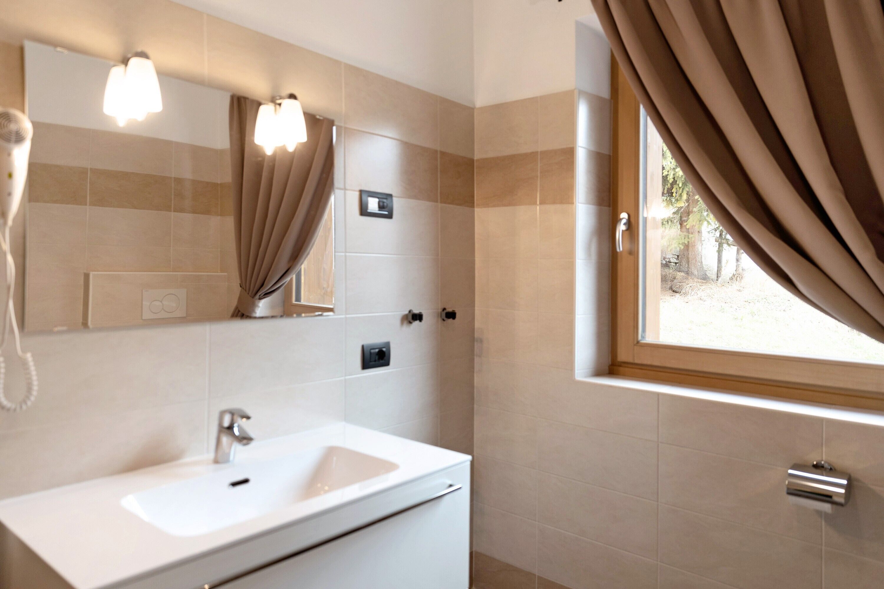 Un baño con un lavamanos blanco, un espejo y un perchero para toallas. Hay una ventana con cortinas y vistas al exterior.