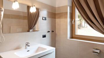 Un baño con un lavamanos blanco, un espejo y un perchero para toallas. Hay una ventana con cortinas y vistas al exterior.
