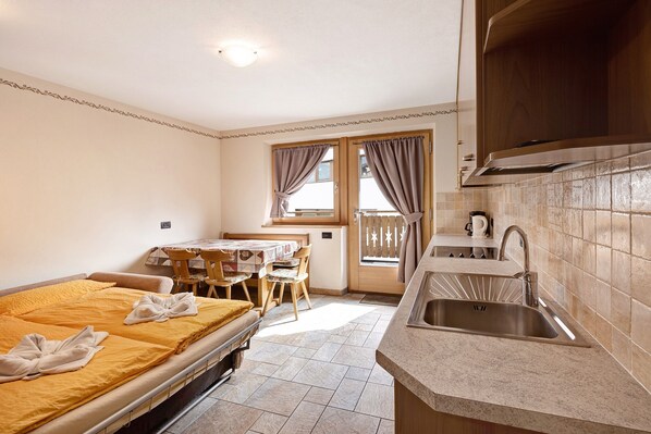 A compact hotel room with a bed, a small dining table, a kitchenette, and a window with curtains.