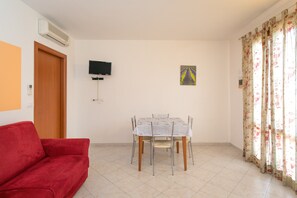 Une pièce avec un canapé rouge, une table à manger entourée de chaises, un téléviseur fixé au mur et une fenêtre ornée de rideaux à motifs floraux.