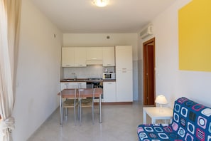 Un espace de vie compact comprenant un coin cuisine, un coin repas avec des chaises, une petite table avec une lampe et un canapé coloré.