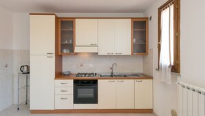 A compact kitchen with a stove, oven, and sink, wooden cabinets, and a window with curtains. - Apartment 'Bilocale Ansonica' with Balcony, Shared Pool & Wi-Fi (Suvereto)