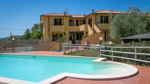 Une piscine avec une terrasse carrelée, un bâtiment à deux étages surmonté d’un toit en tuiles rouges et un espace clos aménagé avec du mobilier d’extérieur.