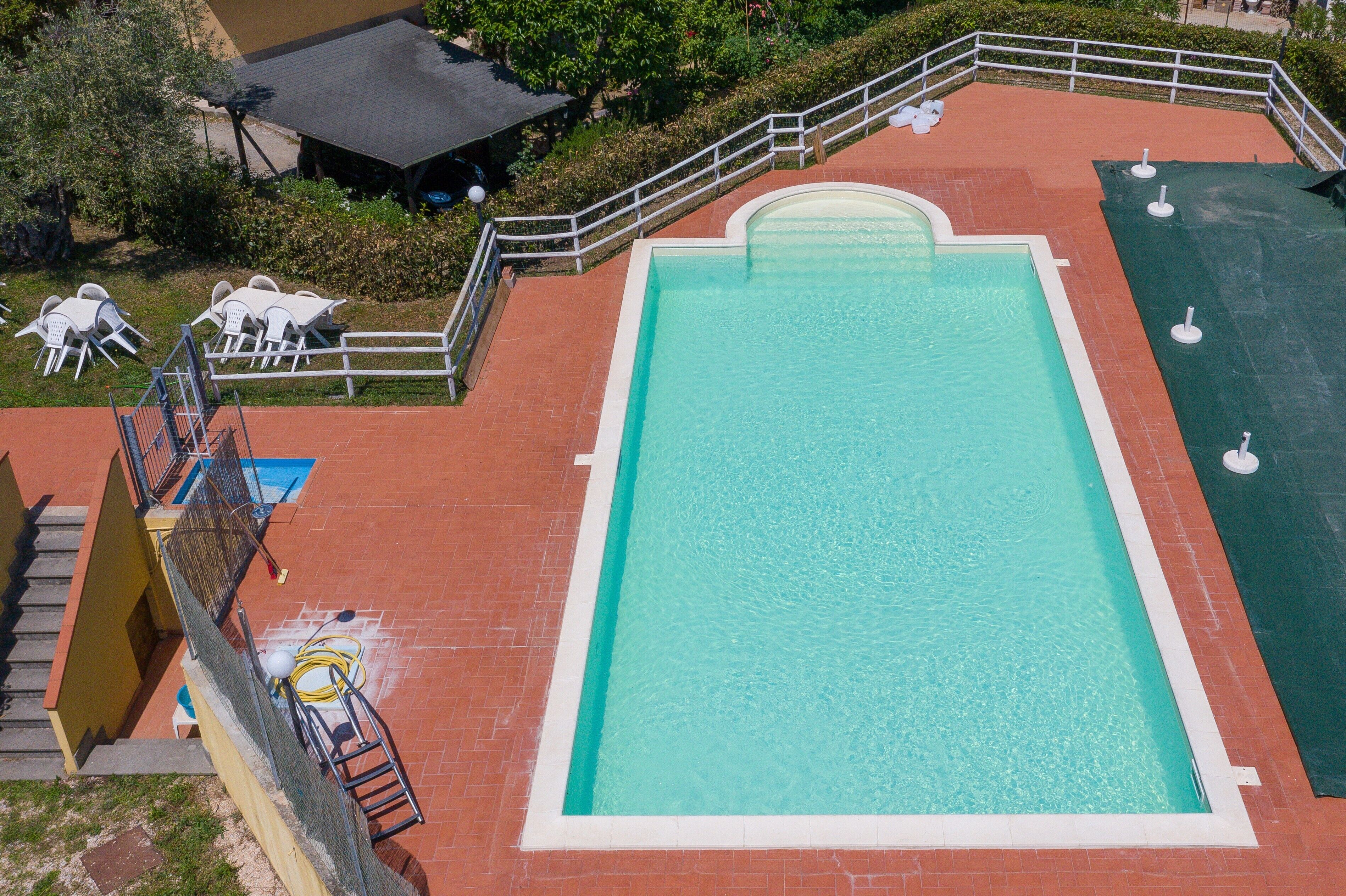 Une piscine rectangulaire bordée de blanc, entourée d’une zone pavée de briques rouges et d’une rambarde blanche.