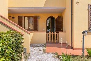 Un bâtiment à la façade jaune, avec des volets en bois, un petit portail et un chemin recouvert de gravier.