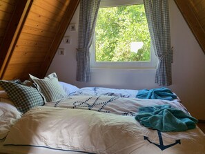 Een gezellige slaapkamer met een schuine kap, een groot raam met gordijnen, een bed met een bedsprei met dessin, en een blauwe deken.