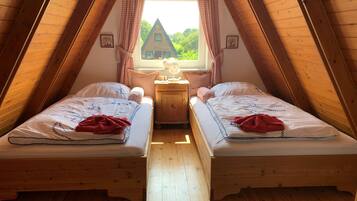 A cozy attic bedroom with two wooden beds, a nightstand, and a window with a view of a house and greenery.