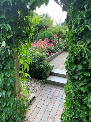 A stone pathway leading through a garden with lush greenery and pink flowers. - Ferienhaus am Meer, Deutschland (Friedrichskoog)