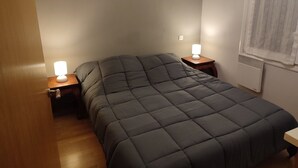 A small, neatly made bed with a dark quilt in a room with wooden flooring and a window with blinds.